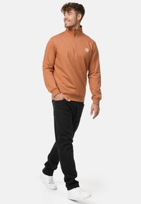 Orange, tekstureret pullover med en kvart lynlås og et logo, kombineret med sorte bukser og hvide sneakers på en model i en neutral baggrund.