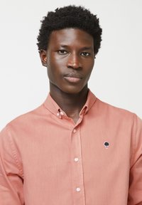 Lichtroze overhemd met knopen en een klassieke kraag, met een kleine boomgeborduurde op de borst en witte knopen. Glad stofmateriaal.