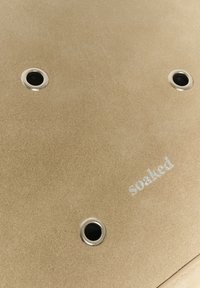 Surface beige avec trois trous circulaires noirs bordés de métal et le mot "soaked" imprimé en petites lettres blanches.