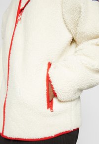 Giacca in pile color crema con cerniera e rifiniture rosse, dotata di tasche laterali con zip e tessuto morbido. Collo rotondo, design casual.