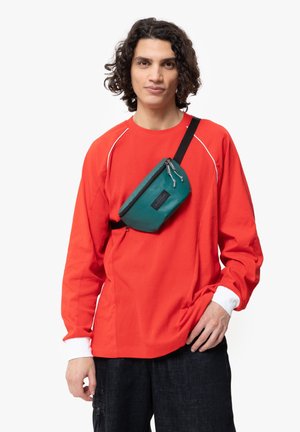 Camicia rossa a maniche lunghe con accenti bianchi, abbinata a una borsa a tracolla verde rettangolare con chiusura zip e superficie testurizzata.