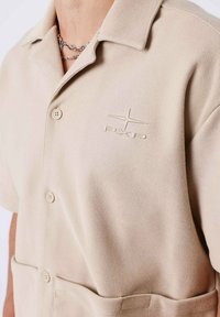 Camicia beige a maniche corte in tessuto strutturato, con colletto, due tasche frontali e un logo ricamato sul petto.