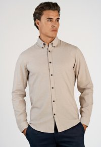 Uomo che indossa una camicia beige con bottoni neri e pantaloni scuri, in piedi con le mani nelle tasche, che guarda di lato contro uno sfondo semplice.