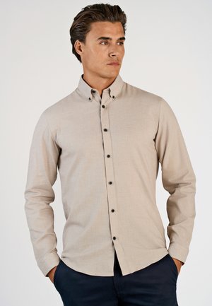 Man in een beige overhemd met zwarte knopen en donkere broek, staand met zijn handen in zijn zakken, kijkend opzij tegen een effen achtergrond.