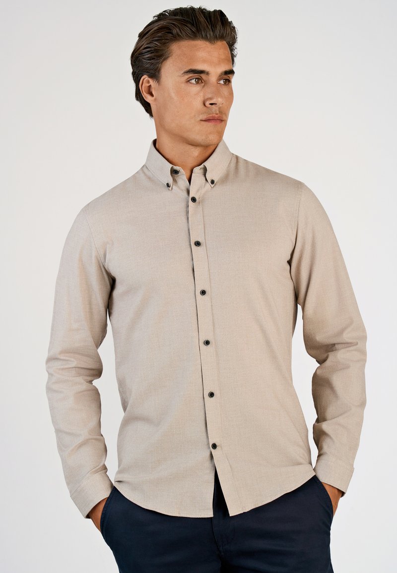 Uomo che indossa una camicia beige con bottoni neri e pantaloni scuri, in piedi con le mani nelle tasche, che guarda di lato contro uno sfondo semplice.