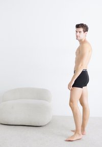 Schwarze Boxershorts an einer männlichen Figur, die seitlich gegen eine weiße Wand steht; im Hintergrund ein plüschiger, hellfarbiger Stuhl. Glatte Hautstruktur sichtbar.