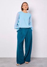 Hellblauer Pullover mit durchsichtigen, karierten Ärmeln, kombiniert mit weit geschnittenen tealblauen Hosen. Sandalen runden den Look ab, mit einem schlichten Ausschnitt.