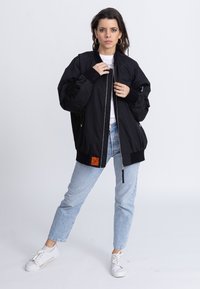 Schwarze Bomberjacke mit einem Reißverschluss vorne, gerippten Bündchen und einem orangefarbenen Etikett. Kombiniert mit hellblauen Jeans und weißen Sneakers.