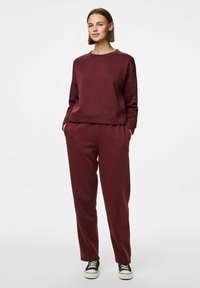 Mulher em pé com as mãos nos bolsos, vestindo um conjunto de sweatshirt e calças burgundy combinando, com ténis pretos e brancos, contra um fundo branco.