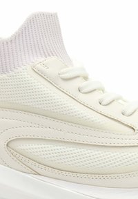 Primer plano de una zapatilla de caña alta de punto blanco y cuero con suela acolchada y cuello texturizado en el tobillo.