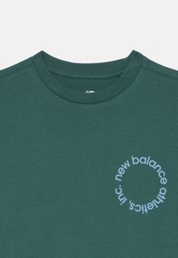 T-shirt de gola redonda verde-escuro com um logo circular azul-claro "new balance athletics, inc." no lado esquerdo do peito.