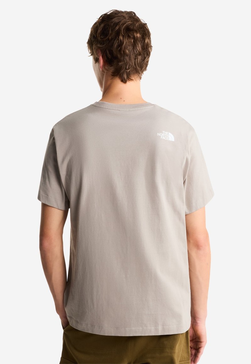 Joven con pelo ondulado que lleva una camiseta gris claro de The North Face, visto desde atrás, de pie frente a un fondo liso.