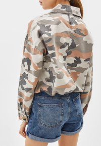 Camicia a bottoni in camo grigia, beige e ruggine, abbinata a pantaloni di jeans corti arrotolati. Presenta un colletto e un dettaglio con piega sul retro.