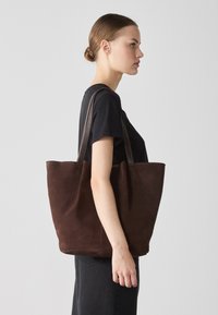 Brown suede tote bag with dual leather straps, open top, and minimalist design. Smooth texture and structured shape for everyday use.