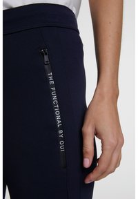 Marinblåbyxor med texturerad duk, en sidoficka med dragkedja märkt med vit text "THE FUNCTIONAL BY OUI" och en figurnära silhuett.