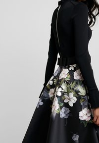Zwarte langemouwen top met een hoge kraag en een rits aan de achterkant, gecombineerd met een rok met bloemenpatroon met grote lichte en donkere bloemen.