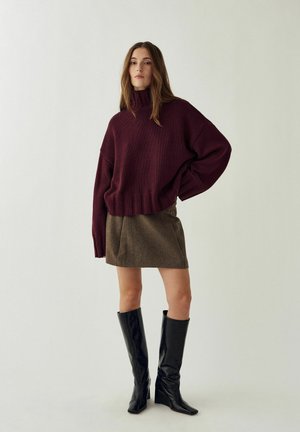 Donna in piedi con maglione a collo alto bordeaux, minigonna marrone e stivali neri al ginocchio su uno sfondo chiaro e semplice.