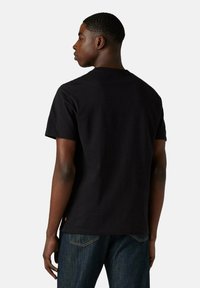 Joven de pie de perfil hacia la izquierda, vistiendo una camiseta negra de manga corta y jeans oscurs de color azul ante un fondo claro y liso.