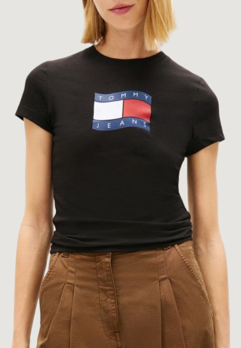 Femme portant un t-shirt noir ajusté avec un logo rectangulaire Tommy Jeans en blanc, rouge et bleu, associé à un pantalon marron taille haute.