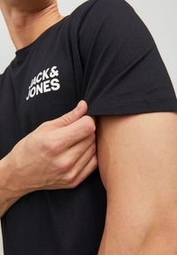 Musta puuvillainen T-paita, jossa on lyhyet hihat ja valkoinen logo vasemmassa yläkulmassa. Kangas on sileäpintainen ja malliltaan klassinen.