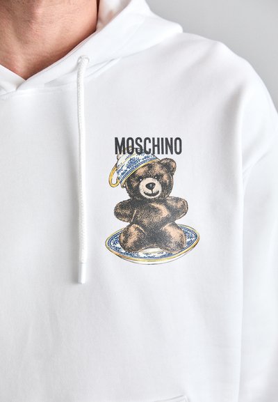 Hanorac alb cu un grafic ce prezintă un urs brun pe o farfurie albastră și albă, purtând o cană decorativă. "Moschino" tipărit deasupra designului.