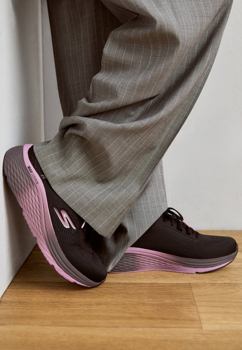 Czarne buty do biegania z teksturowaną cholewką, grubą różową podeszwą środkową i czarnymi sznurowadłami, zestawione z szarymi spodniami w prążki. Stojące przy ścianie.