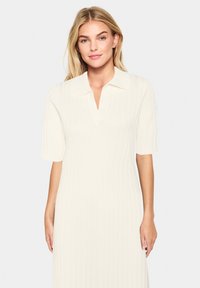 Robe jusqu'aux genoux en tricot côtelé crème, dotée de manches courtes et d'un col avec une découpe en V subtile. Texture lisse et extensible.