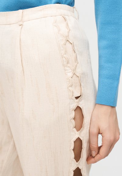 Μπεζ παντελόνι από λινό με κυματιστές ανοίγματα κατά μήκος του εξωτερικού μηρού, φορεμένο με μπλε μπλούζα μακρύ μανίκι και εμφανές χέρι.