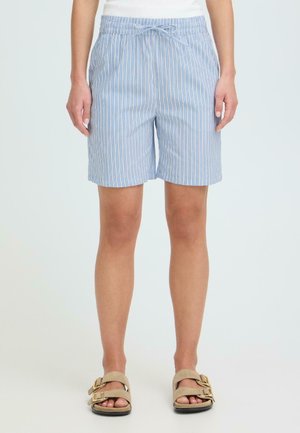 Kvinde iført lyseblå og hvide, knælange shorts med elastisk taljebånd og beige sandaler med spænde, stående på hvid baggrund.