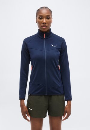 Γυναίκα που φοράει σκούρο μπλε τζάκετ με φερμουάρ και πορτοκαλί τράβηγμα φερμουάρ και χακί πράσινα σορτς, στέκεται μπροστά σε απλό φωτεινό φόντο.