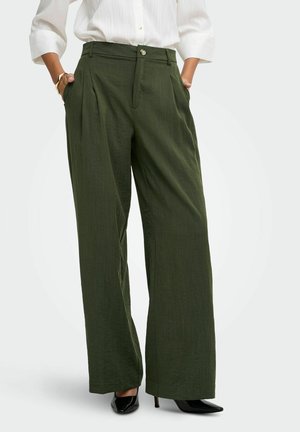 Femme portant un pantalon taille haute, large, vert olive avec des plis, associé à des talons pointus noirs et une chemise blanche boutonnée aux manches trois-quarts.
