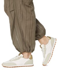 Sneaker bianchi e beige con un design testurizzato, dettagli dorati e suola in gomma. Abbinati a pantaloni larghi color verde oliva.