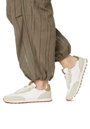 White and beige sneakers with a textured design, gold accents, and a rubber sole. Paired with loose, olive green pants.