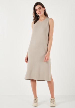Sleeveless midi dress in light beige, made from soft fabric with a relaxed fit and side slits, paired with light-colored sneakers.