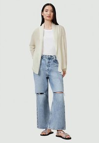 Cardigã de creme claro sobre uma blusa branca, combinado com jeans azul de perna larga desgastados e sandálias pretas. Textura de malha suave, ajuste descontraído.