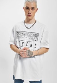 Hvit oversized t-skjorte med svart paisley-design og ordet "LEGEND" i fet skrift, kombinert med blå jeans og en sølvkjede halskjede.