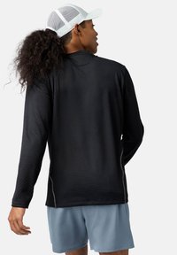 Langarm schwarzes Sportshirt aus strukturiertem Stoff, mit Seitennähten und reflektierenden Akzenten, kombiniert mit hellblauen Shorts und einer weißen Mütze.