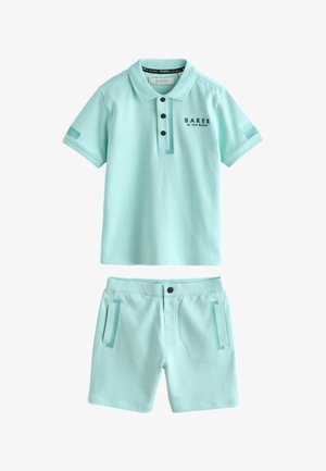 Jongen draagt een lichtblauw poloshirt en bijpassende korte broek, staat tegen een muur met handen in de zakken en witte sneakers.