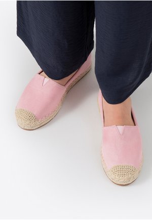 Füße tragen pinkfarbene Wildleder-Espadrilles mit geflochtenen Zehenkappen und marineblaue Schlaghosen vor weißem Hintergrund.