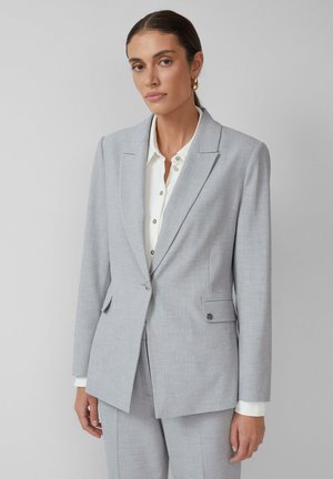 Lichtgrijze op maat gemaakte blazer met een gestructureerd ontwerp, reverskraag, twee voorzakken en een enkele knoopsluiting boven een wit shirt.