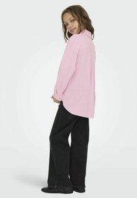 Chemise rose clair surdimensionnée avec des poignets à boutons, associée à un jean large noir et des chaussures noires. Design simple et décontracté.