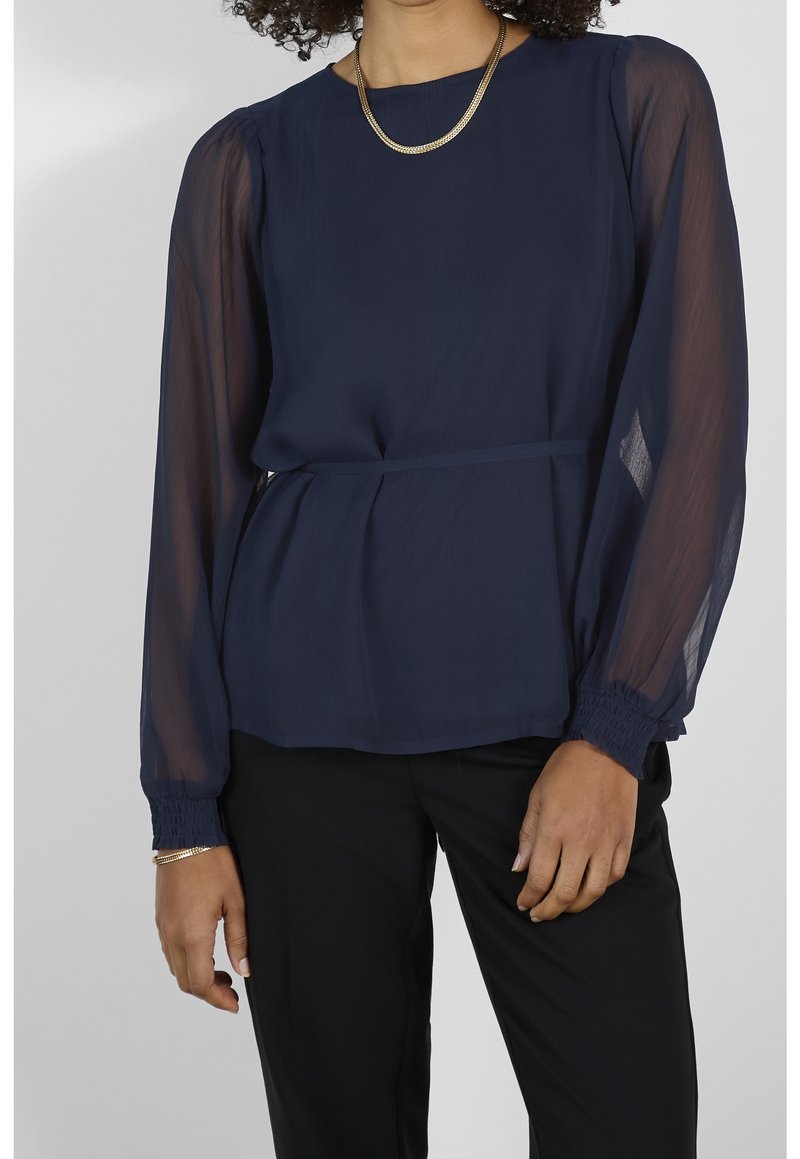 Femme portant un chemisier bleu marine aux manches longues transparentes, assorti à un pantalon noir ainsi qu'un collier et un bracelet en or.