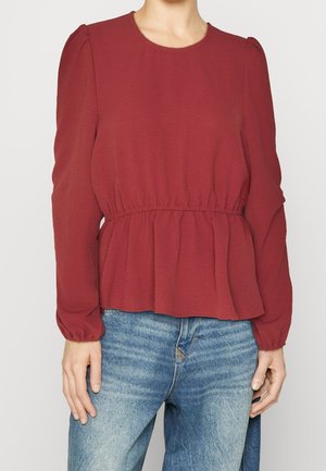 Vrouw draagt een lange mouwen rode blouse met elastische taille en blauwe jeans, staand tegen een effen lichte achtergrond.
