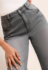 Kõrge vöökohaga hallid denim teksad nööbiga sulgemisega, ees taskud ja sile, tekstureeritud viimistlus. Käsi asetatud reie piirkonda.