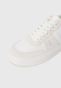 Λευκά αθλητικά παπούτσια από σουέτ και καμβά με κρεμ χερού λια, διακοσμημένα με ασημένιο λογότυπο και υφή στην σόλα. Μοντέρνος, μινιμαλιστικός σχεδιασμός.