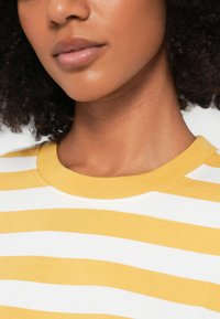 Κίτρινο και λευκό ριγέ βαμβακερό μπλουζάκι με στρογγυλή λαιμόκοψη και λεία υφή· κοντινή εικόνα που τονίζει το γιακά και τις λεπτομέρειες των ραφών.