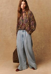Top a maniche lunghe con motivo floreale e scollatura con fiocco marrone, abbinato a pantaloni in denim a gamba larga di colore azzurro chiaro. Borsa marrone con dettaglio ad anello.