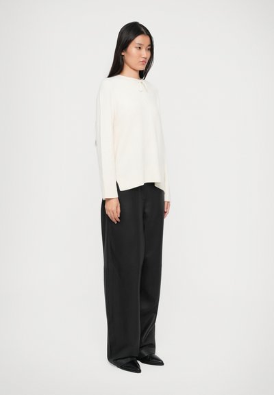 Light cream sweater with a front tie detail, paired with loose black trousers and pointed black shoes. Simple, relaxed fit design.