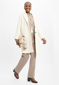 Crèmekleurige jas met een op maat gemaakt ontwerp, gecombineerd met een beige outfit en bruine haklaarzen. Houdt een gewatteerde lichtbeige tas vast.