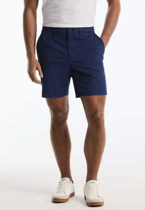 Mann trägt marineblaue maßgeschneiderte Shorts, weiße Turnschuhe und ein weißes T-Shirt, steht mit einer Hand in der Tasche.